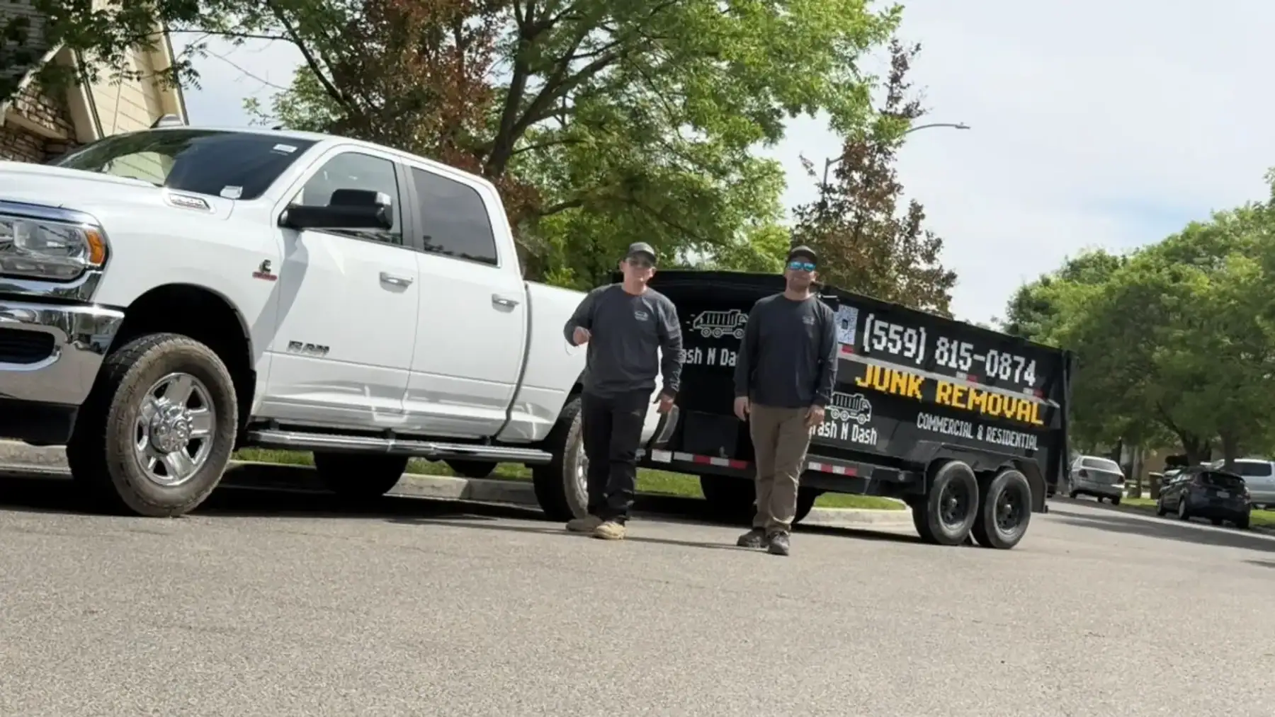 Two Trash N Dash crew members standing beside a white pickup and the black Trash N Dash dump trailer on a street