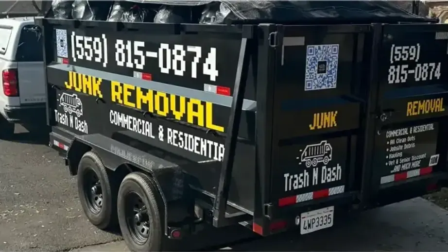 The black Trash N Dash dump trailer with yellow Junk Removal lettering and the phone number on its side